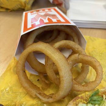 Whataburger - Onion Rings - Golden-brown onion rings are placed in a paper container on a yellow napkin, with a burger and other food items partially visible. - Fast Food Near Me - Phoenix, Arizona