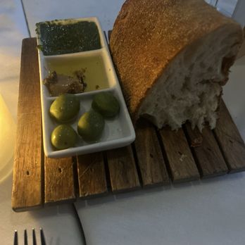 Assaggio - A large loaf of bread is placed on a wooden serving board, accompanied by three small dishes containing sauces and four green olives. - Italian Near Me - Boston, Massachusetts