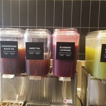 CAVA - The juice machine - Four large beverage dispensers with labels for different drinks are positioned on a counter, with a black tiled wall in the background. - Mediterranean Near Me - Cedar Park, Texas