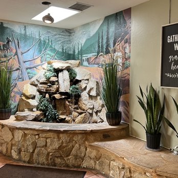 Casa De Benavidez - a fountain in the lobby - An indoor water feature composed of stacked rocks with small plants is situated on a stone base. The background features a mural of a mountain landscape with trees and a waterfall. Several potted plants are placed around the water feature, and a decorative sign with the message "Gather here with grateful hearts" hangs on the wall. The space has tiled flooring and ceiling lighting. - Mexican Near Me - Albuquerque, New Mexico