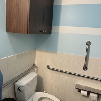Poke Wave - Restroom - A public restroom features a white toilet, beige tiled walls, and blue painted walls with white stripes. Wall-mounted cabinets and safety grab bars are visible. - Poke Near Me - Tempe, Arizona