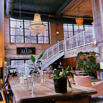 The Allis - Indoor - A spacious restaurant interior with large windows, chandeliers, potted plants, and a staircase leading to an upper level. - Lounges Near Me - Chicago, Illinois