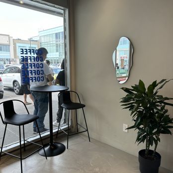 A Lot Coffee - Interior of a coffee shop featuring a window with people outside, two black chairs, a small round table, a decorative mirror, and a potted plant. - Coffee & Tea Near Me - Markham, Ontario