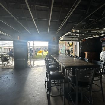 Celis Brewery - Inside - The image shows the interior of a brewery with a dark ceiling and high tables with chairs. Open doors at the back reveal an outdoor area with people, vehicles, and a grassy space. The lighting inside is dim, with natural light coming from outside. The space appears to be a casual dining or drinking area. - Breweries Near Me - Austin, Texas