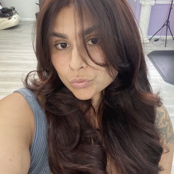 Sunshine Studio - A woman with long, wavy brown hair and a nose piercing is taking a selfie in a salon. The background includes light-colored flooring, purple walls, a tripod, and salon equipment. She is wearing a sleeveless top and has a tattoo on her shoulder. - Makeup Artists Near Me - Arlington, Virginia