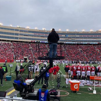 Camp Randall Stadium - ESPN at work - A cameraman stands on a raised platform filming a football game at Camp Randall Stadium, with players on the field and a large crowd in the stands. - Stadiums & Arenas Near Me - Madison, Wisconsin
