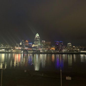 Beeline - Skyline view from beeline - Night scene of Cincinnati's downtown skyline featuring illuminated skyscrapers, including a prominent tall building with a rounded top. The city lights reflect on the calm Ohio River, and the sky is dark with no visible stars. The foreground shows a riverbank with minimal features, emphasizing the cityscape and its reflections. - Bars Near Me - Newport, Kentucky
