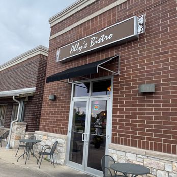 Ally's Bistro - entrance - The photo depicts the front exterior of Ally's Bistro, a casual American restaurant. The building has a brick facade with a stone base, and a sign displaying the restaurant's name is mounted above the entrance. There is a black awning over the glass door, which has a neon sign and menu details visible through the window. Outside, there are three small round tables with chairs arranged on the sidewalk, providing outdoor seating. The sky appears overcast, and the area looks clean and well-maintained. - Cafes Near Me - Menomonee Falls, Wisconsin
