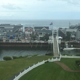 Bridge to Perkins cove - The image shows a coastal neighborhood with a row of houses facing the water, featuring various colors and architectural styles. A dock extends into the water with a small boat moored nearby. In the foreground, there is a well-maintained grassy area with a white fence and benches. The ocean and a cloudy sky are visible in the background, indicating a seaside setting.
