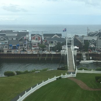 Auberge On The Cove - Bridge to Perkins cove - The image shows a coastal neighborhood with a row of houses facing the water, featuring various colors and architectural styles. A dock extends into the water with a small boat moored nearby. In the foreground, there is a well-maintained grassy area with a white fence and benches. The ocean and a cloudy sky are visible in the background, indicating a seaside setting. - Hotels Near Me - Ogunquit, Maine