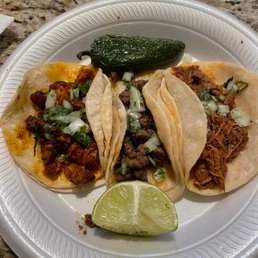 Pastor - A disposable plate holds three tacos with various fillings: one with chopped meat and vegetables, another with shredded meat, and the third with seasoned meat, all topped with chopped onions and herbs. A lime wedge and a roasted pepper are also on the plate, set on a granite countertop.