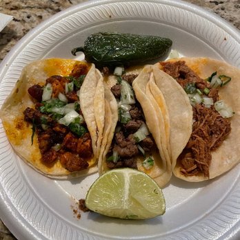 Mad Taco AZ - Al pastor, Carne asada, Birria - A disposable plate holds three tacos with various fillings: one with chopped meat and vegetables, another with shredded meat, and the third with seasoned meat, all topped with chopped onions and herbs. A lime wedge and a roasted pepper are also on the plate, set on a granite countertop. - Food Trucks Near Me - Apache Junction, Arizona