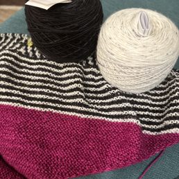 Knitting project and yarn. - The image shows two yarn balls, one black and one white, resting on a knitted fabric with black and white horizontal stripes and a pink section. The yarn balls are placed on a teal-colored surface, with the black yarn ball having a paper label and the white yarn ball having a rolled paper inside. The knitted fabric appears to be part of a knitting project, with the yarn balls ready for use.