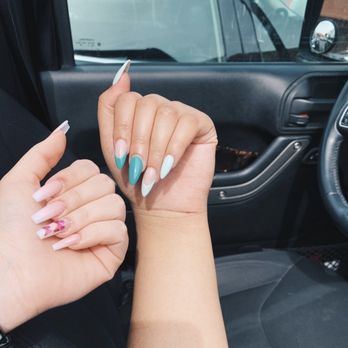 Famous Nails - Two hands with manicured nails are shown inside a vehicle, with one hand displaying a pink and floral design and the other featuring a blue and white gradient. The background includes the vehicle's dashboard, steering wheel, and side mirror, indicating the photo was taken inside a car. - Nail Salons Near Me - Albuquerque, New Mexico
