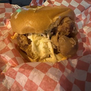 PG Clucks - A fried chicken sandwich features a crispy fried chicken fillet topped with melted cheese, served in a soft bun. The sandwich is wrapped in red and white checkered paper, placed on a table, with a background suggesting an outdoor or casual dining setting. - Sandwiches Near Me - Toronto, Ontario