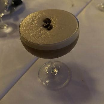 Bacaro - Espresso martini - A photograph of an espresso martini served in a coupe glass, featuring a smooth, creamy top layer garnished with coffee beans. The drink is placed on a table with dim lighting, and the background includes blurred tableware and glassware, indicating a restaurant or bar setting. - Wine Bars Near Me - Providence, Rhode Island
