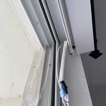 MDDesigns - The image depicts a close-up view of a window frame, showing aluminum tracks, a white cable, and hardware components. The window is set within a wall with textured white paint, and the wiring appears to be exposed, possibly indicating ongoing installation or maintenance work. The focus is on the structural and electrical elements around the window area. - Shades & Blinds Near Me - Miami, Florida