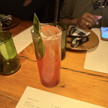 Basso - A tall, clear glass contains a pink cocktail with ice, garnished with a green leaf and a black straw. The drink is on a wooden table alongside menus, glasses, and a plate with a napkin. Two people are seated at the table, one using a smartphone, in a dimly lit restaurant setting. - Bars Near Me - Saint Louis, Missouri