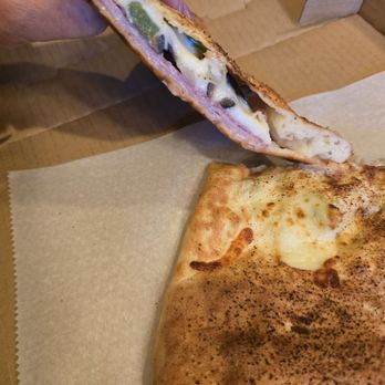 Davidos - Italian Stromboli - A person holds a slice of pizza with toppings including cheese, jalapeños, and mushrooms, showing the cross-section of the slice. The pizza has a golden-brown crust and is placed on parchment paper on a wooden surface. The slice reveals melted cheese and toppings inside, with the remaining pizza visible in the foreground. - Pizza Near Me - Rio Rancho, New Mexico