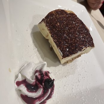 Rustico Taverna - Tiramisu - A rectangular slice of tiramisu with a cocoa powder dusted top is placed on a white square plate. Next to it, there is a dollop of whipped cream topped with berry sauce and a cherry. The plate has some powdered sugar sprinkled around the dessert. A person's hand with red nail polish is visible in the background. - Italian Near Me - Queens, New York