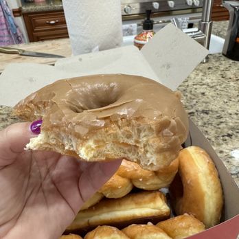 Rancho Donuts - The most delicious maple glaze! - A hand holds a glazed donut with a bite taken out of it, above a box of various donuts on a kitchen countertop. - Donuts Near Me - Temecula, California