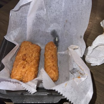 Twisted Wings  - MOZZARELLA (5) STICKS - Two fried mozzarella sticks are placed on parchment paper inside a black plastic basket, with some crumbs and a crumpled napkin nearby. - Chicken Wings Near Me - Indianapolis, Indiana
