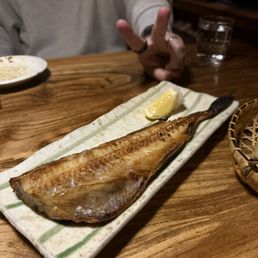 A grilled fish fillet served on a rectangular ceramic plate with a lemon wedge, on a wooden table in a restaurant setting.