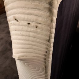 A close-up image of a wooden furniture leg, displaying natural wood grain patterns, knots, and a smooth, sanded surface. The leg is attached to a dark-colored piece of furniture, possibly a table or cabinet, with a focus on the natural texture of the wood.