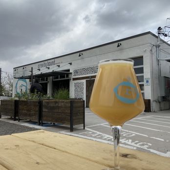 Gravity Bound Brewing - A tulip-shaped glass filled with beer is positioned on a wooden table outdoors. The background features the exterior of Gravity Bound Brewing, a brewery in Albuquerque, NM, with a modern, minimalist facade, some greenery, and a cloudy sky overhead. The scene suggests a casual outdoor setting for enjoying craft beer. - Brewpubs Near Me - Albuquerque, New Mexico