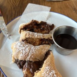 A white plate holds several pieces of fried pastry with a dusting of powdered sugar, filled with chocolate and possibly nuts. A small metal cup containing dark syrup is placed on the plate. The plate is on a wooden table, with a knife and a fork visible nearby.