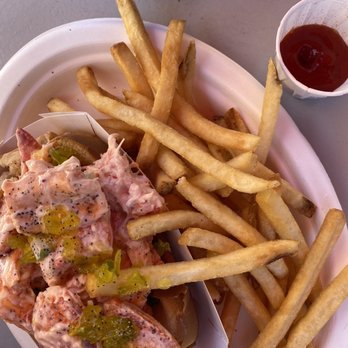 J T's Seafood - Lobster salad roll with fries - A food tray contains a lobster salad roll with chopped celery and pickles, served in a bun. French fries are piled next to the roll. A small cup of ketchup and a Pepsi soft drink are also on the table. The tray is on a gray surface, and the Pepsi cup is partially visible at the top of the image. - Seafood Near Me - Brewster, Massachusetts