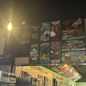 Launch Pad Brewery - Beer wall - A set of colorful illustrated menus are mounted on a wall, featuring craft beer names and descriptions with space and holiday themes. The artwork includes astronauts, Christmas imagery, and winter scenes, with some menus highlighting specific flavors like barrel-aged barleywine and hot cocoa. The menus are arranged above a bar area with visible refrigerators and framed pictures, and the lighting includes a hanging bulb and neon accents. - Breweries Near Me - Aurora, Colorado