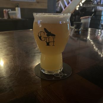 Ghost Hawk Brewing - Apex predator IPA - A glass of beer with a logo, containing a light-colored beverage, is placed on a dark wooden bar table in a dimly lit setting. - Breweries Near Me - Clifton, New Jersey