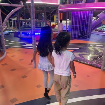 Flying Apple - Two girls walk through an indoor playground area with vibrant, colorful play structures and lighting, with other children and adults in the background. - Indoor Playcenter Near Me - Brooklyn, New York