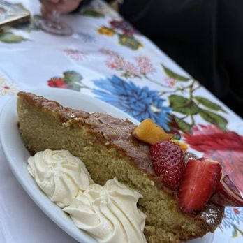 Bombera - A slice of cake with whipped cream and fresh strawberries is served on a white dish on a floral tablecloth, with a person holding a drink in the background. - Mexican Near Me - Oakland, California
