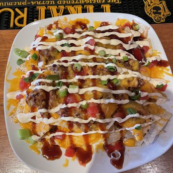Old 97 Cafe - BBQ brisket nachos - A white plate holds a serving of loaded nachos with tortilla chips covered in melted cheese, seasoned meat, and drizzled with sour cream and barbecue sauce. Small pieces of chopped green onions and diced tomatoes are scattered on top. The dish is placed on a wooden table, with a bar mat visible in the background, indicating a bar or restaurant setting. - Jazz & Blues Near Me - Akron, Ohio
