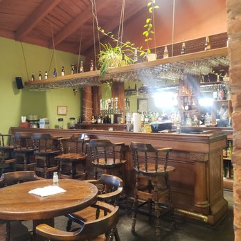 Casa De Valdez - Bar - A wooden bar with stools is set against a green wall, with a hanging shelf above holding bottles and plants, and a mirror behind the bar. - Barbeque Near Me - Taos, New Mexico
