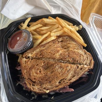 Legacy Table - Ruben - A meal in a black plastic takeout container includes a sandwich with toasted bread, French fries, and a small container of ketchup. The sandwich appears to contain meat and possibly cheese or other fillings. The container is placed on a surface with a plastic bag underneath, and part of a person's hand is visible holding a napkin or paper towel. - Salad Near Me - Kalamazoo, Michigan