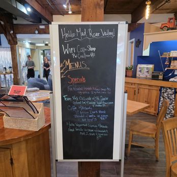 Three Mountain Cafe - Menu - A chalkboard menu in a coffee shop features handwritten text listing specials and Wi-Fi details. The board is positioned on a wooden post inside a space with wooden beams and warm lighting. The menu includes food and drink options, with some items written in different colors for emphasis. The background shows part of the cafe interior, including seating and decor elements. - Sandwiches Near Me - Waitsfield, Vermont