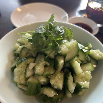 Dumpling Wei - Garlic Cucumber - A white bowl contains a cucumber salad made of chopped cucumbers and garnished with fresh cilantro leaves. The salad appears to be lightly dressed or marinated. In the background, there is a small dish of soy sauce, a glass of dark soda, and an empty plate. The setting suggests a casual dining environment, possibly at a restaurant specializing in Asian cuisine. - Chinese Near Me - Pasadena, California