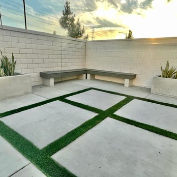 Ultimate Design & Construction - Backyard - An outdoor patio features concrete paving with grass borders, concrete planters with plants, and a concrete bench along the wall, enclosed by a brick wall. - General Contractors Near Me - Riverside, California