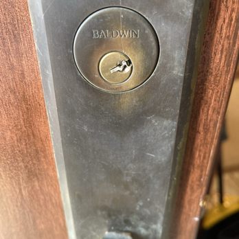 Locksmith Dash - Key extraction service needed - A close-up image of a Baldwin lock installed on a wooden door. The lock features a circular keyhole with visible damage, including a broken or bent key slot. The lock is mounted on a metal plate, which appears slightly worn, and the door frame is made of wood with a visible grain pattern. The image focuses on the lock and the immediate surrounding area, highlighting the damage to the keyhole. - Keys & Locksmiths Near Me