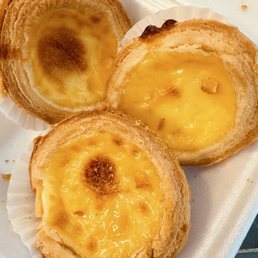 Egg tarts - Three Portuguese egg tarts with flaky, layered pastry crust and smooth, yellow custard filling. The tarts are placed on a foam tray, with some showing slightly burnt spots on the custard surface. The image captures the baked goods in a close-up view, highlighting the texture of the crust and the glossy appearance of the custard.