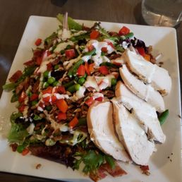 The Farm salad w chicken - A square white plate holds a salad with sliced cooked chicken breast, mixed greens, chopped red bell peppers, green peas, and green beans. The salad is topped with a creamy dressing and small pieces of chopped radish. A glass of water is visible in the background, and the plate is placed on a dark wooden table.