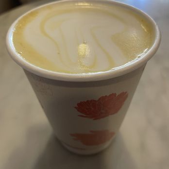 Hibiscus Brew - A paper cup containing a creamy beverage with a yellow and white swirl pattern on the surface, placed on a light-colored table. - Cafes Near Me - Brooklyn, New York