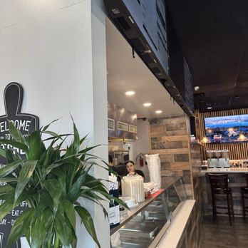 Cuna Del Sol - The image shows a restaurant interior with a counter, a large green plant, and a woman working behind the counter. There are digital screens above and a TV on the wall. - Mexican Near Me - Garnerville, New York