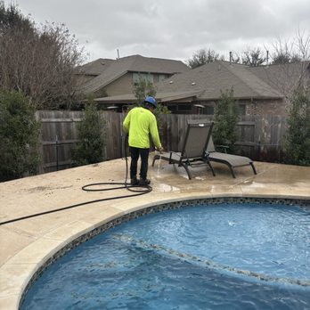 Moon Valley Nurseries - Working hard for final presentation!! - A worker dressed in a neon yellow long-sleeve shirt and blue safety helmet is using a hose to clean the pool deck area. The backyard features a swimming pool with a tiled edge, lounge chairs, and a wooden fence with trees and houses in the background. The sky is overcast, indicating cloudy weather. - Nurseries & Gardening Near Me - Buda, Texas