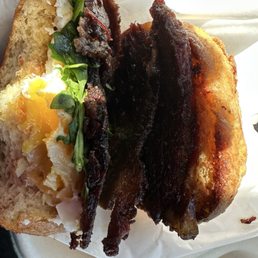 Brisket Buckaroo - A breakfast sandwich featuring fried egg, crispy bacon, and fresh greens on toasted bread, served in a paper wrapper.