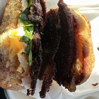 Moore Bread - Brisket Buckaroo - A breakfast sandwich featuring fried egg, crispy bacon, and fresh greens on toasted bread, served in a paper wrapper. - Bakeries Near Me - Denver, Colorado