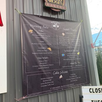 Bread & Roses - Menu! - The image shows a large black menu board mounted on a gray metal wall outside Bread & Roses Bakery. The menu lists various baked goods, including morning bakes like muffins and scones, sweet treats such as cookies and brownies, and cake slices with different flavors. The board also mentions daily changing varieties and available 6-inch cakes. A bakery sign with the name and logo is visible above the menu. - Coffee & Tea Near Me - Wells, Maine